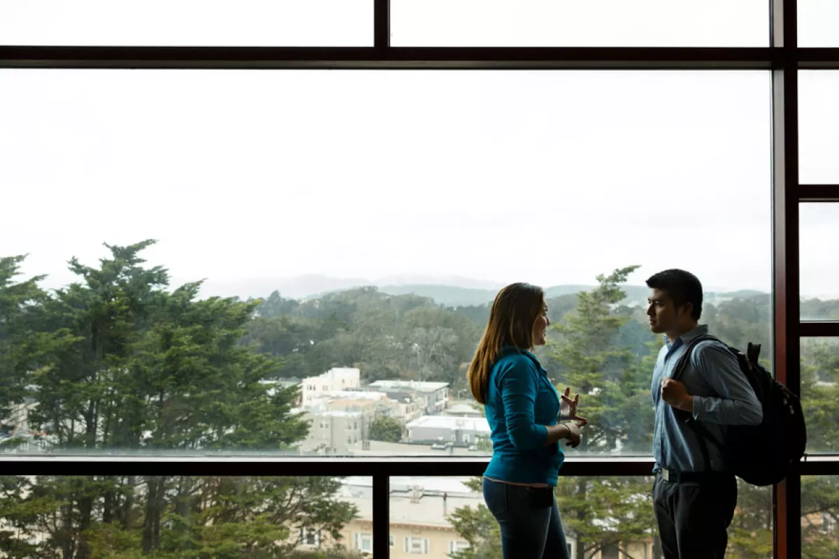 Two UCSF community members confer while standing in front of a wall-to-wall, floor-to-ceiling window in the main library with the the Parnassus campus seen through the widow