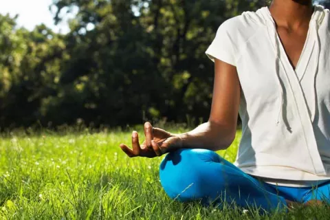 A women sitting in a yoga lotus position on a grass lawn with wooded features in the background